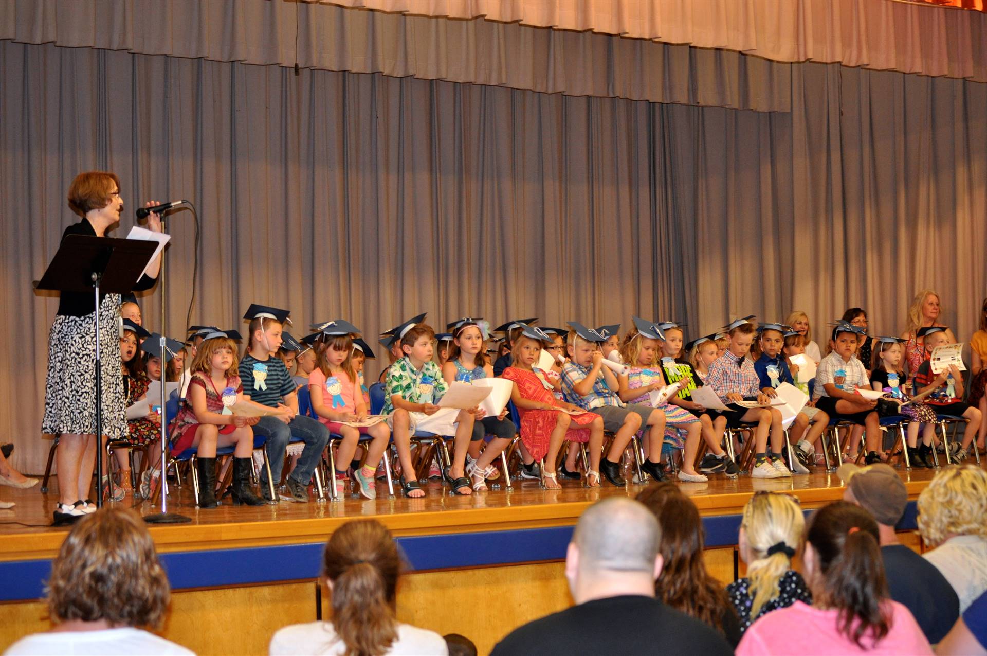 A teacher speaks on stage about 2 special helpers while students listen.