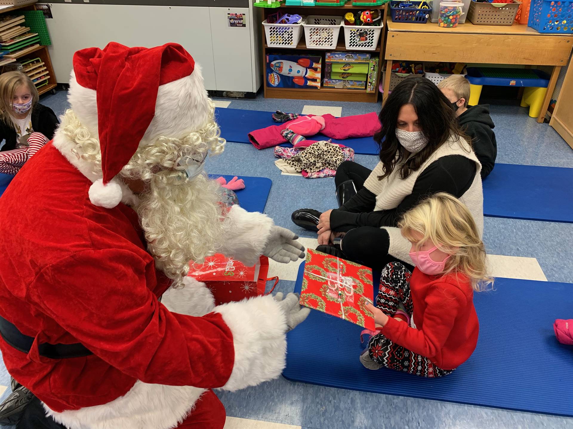 Santa gives a gift to a student while the teacher watches.