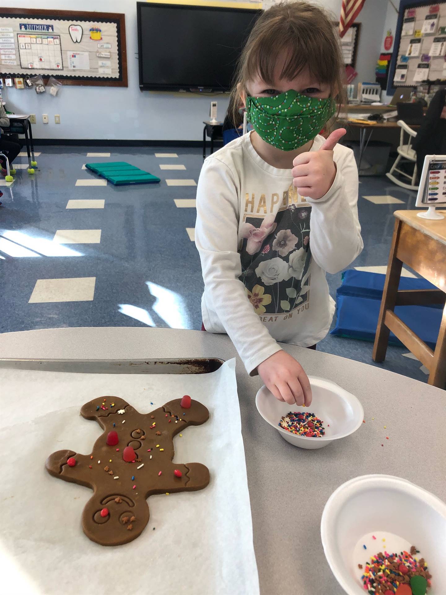 child gives a thumbs up as she adds some sprinkles to her gingerbread cookie