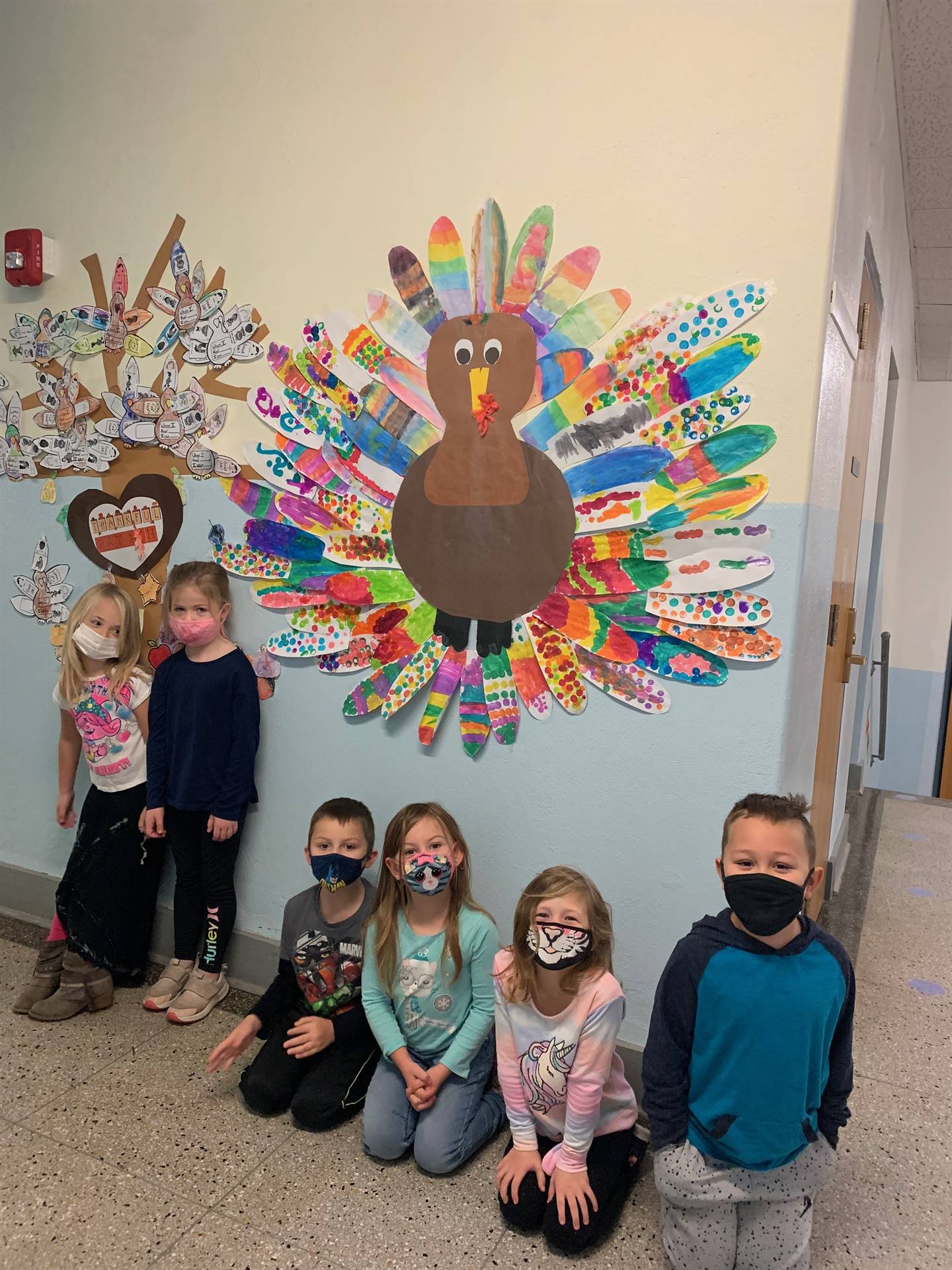 6 students standing in front of a paper turkey with multicolored feathers.