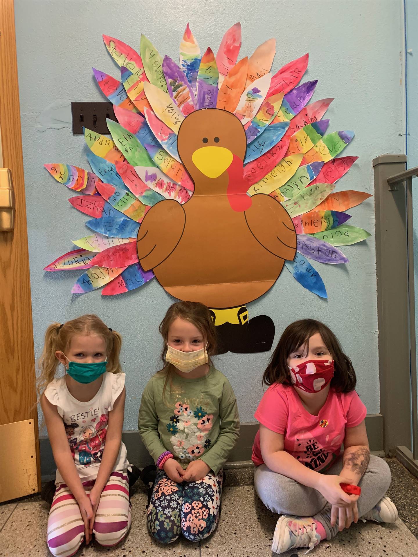 3 students sitting in front of giant paper turkey with multicolored feathers.
