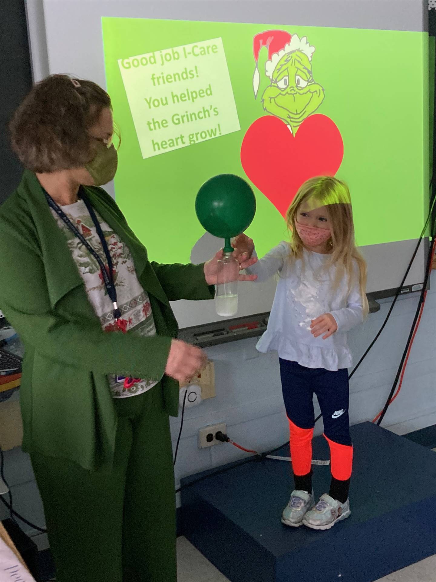 adult and student holding green balloon with grinch face and heart in background.