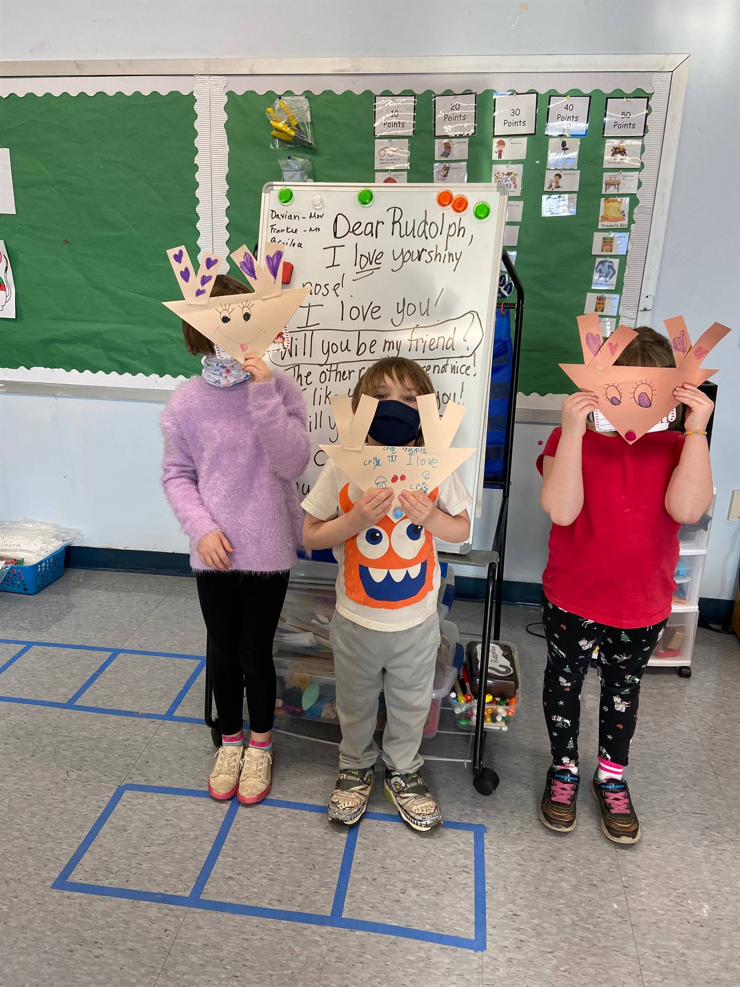 3 students holding paper reindeer heads in front of faces with letter to rudolph  in background 