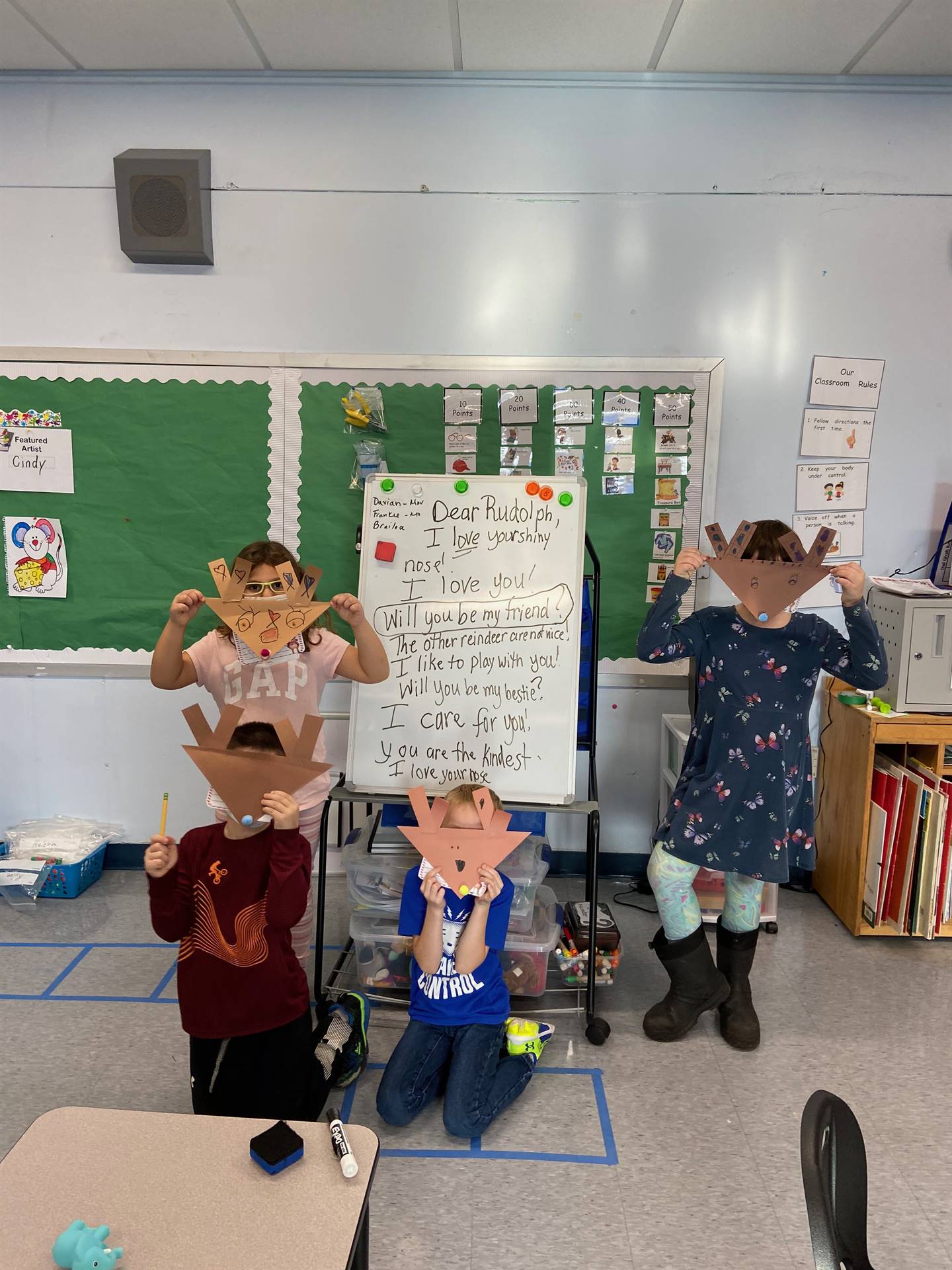 4 students holding paper reindeer heads in front of faces with letter to rudolph  in background 