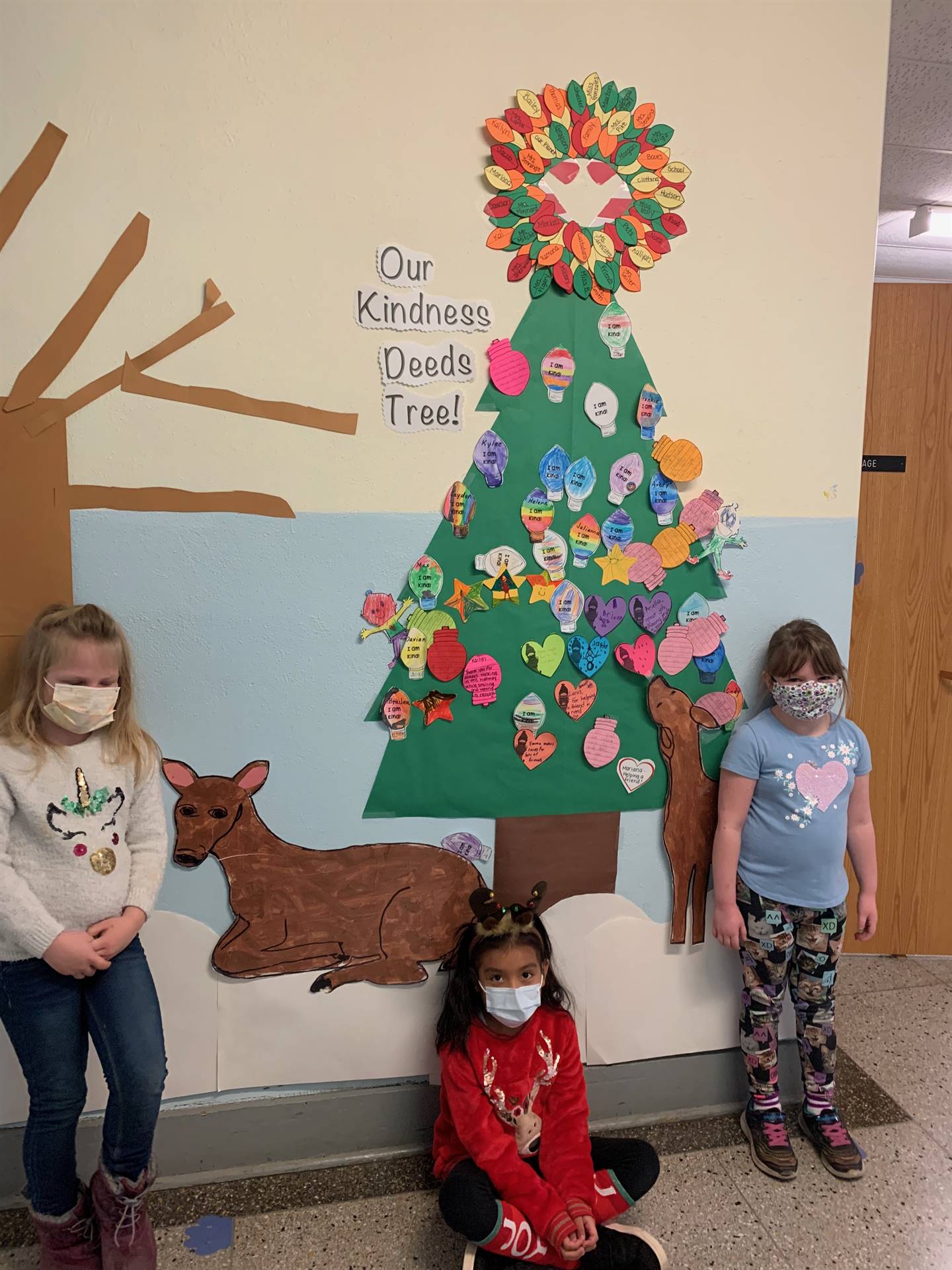 3 students with a paper evergreen tree topped with a light bulb wreath and covered in various colore