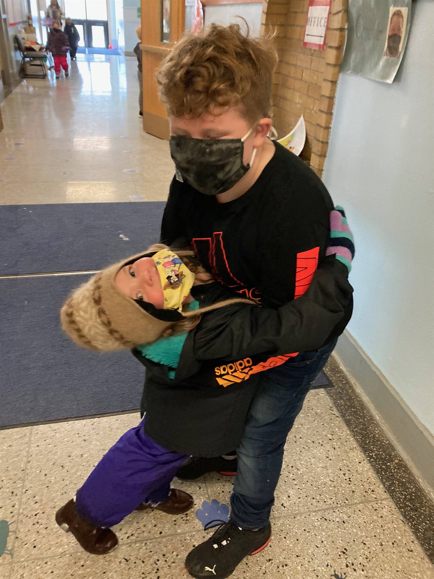 2 students with winter clothes and mask are hugging in a hallway.