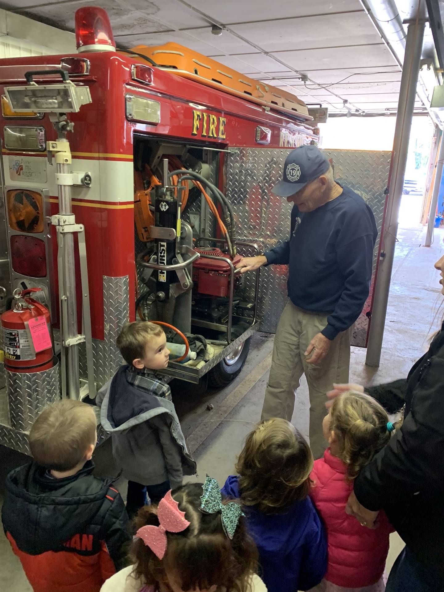 firefighter in hat shows a group of students tools on a firetruck.