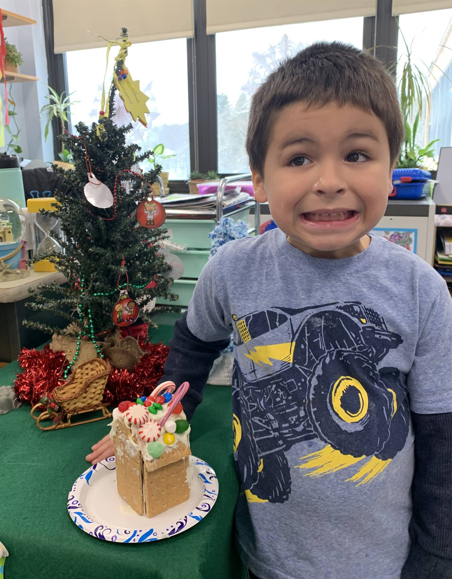 a student standing in front of a decorated tree holding a decorated gingerbread house