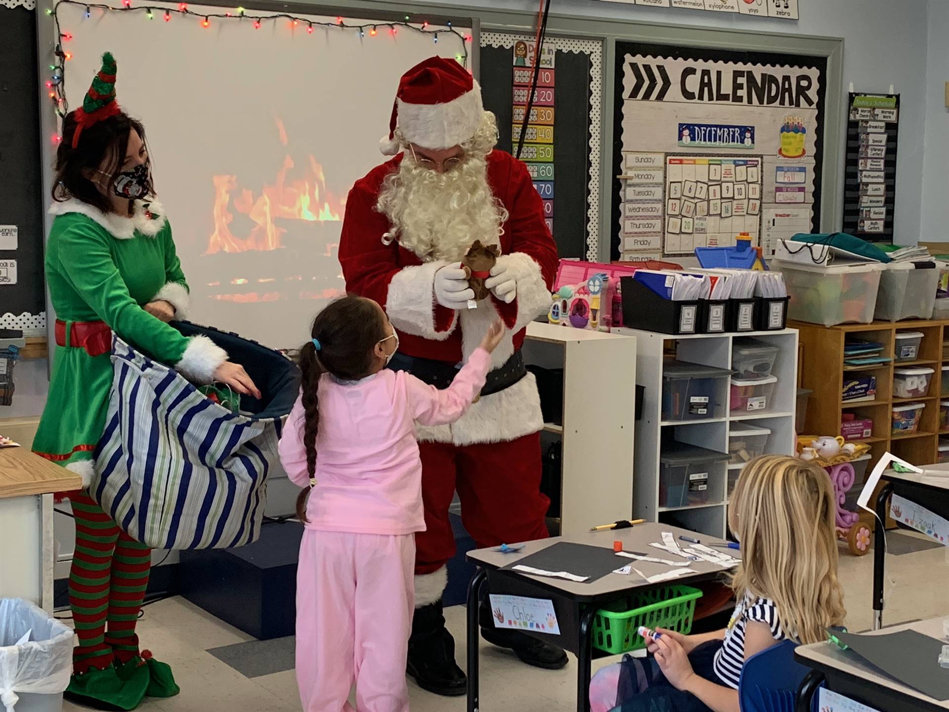 A student hands a present to Santa. Elf stands by and watches in green suit and hat. 