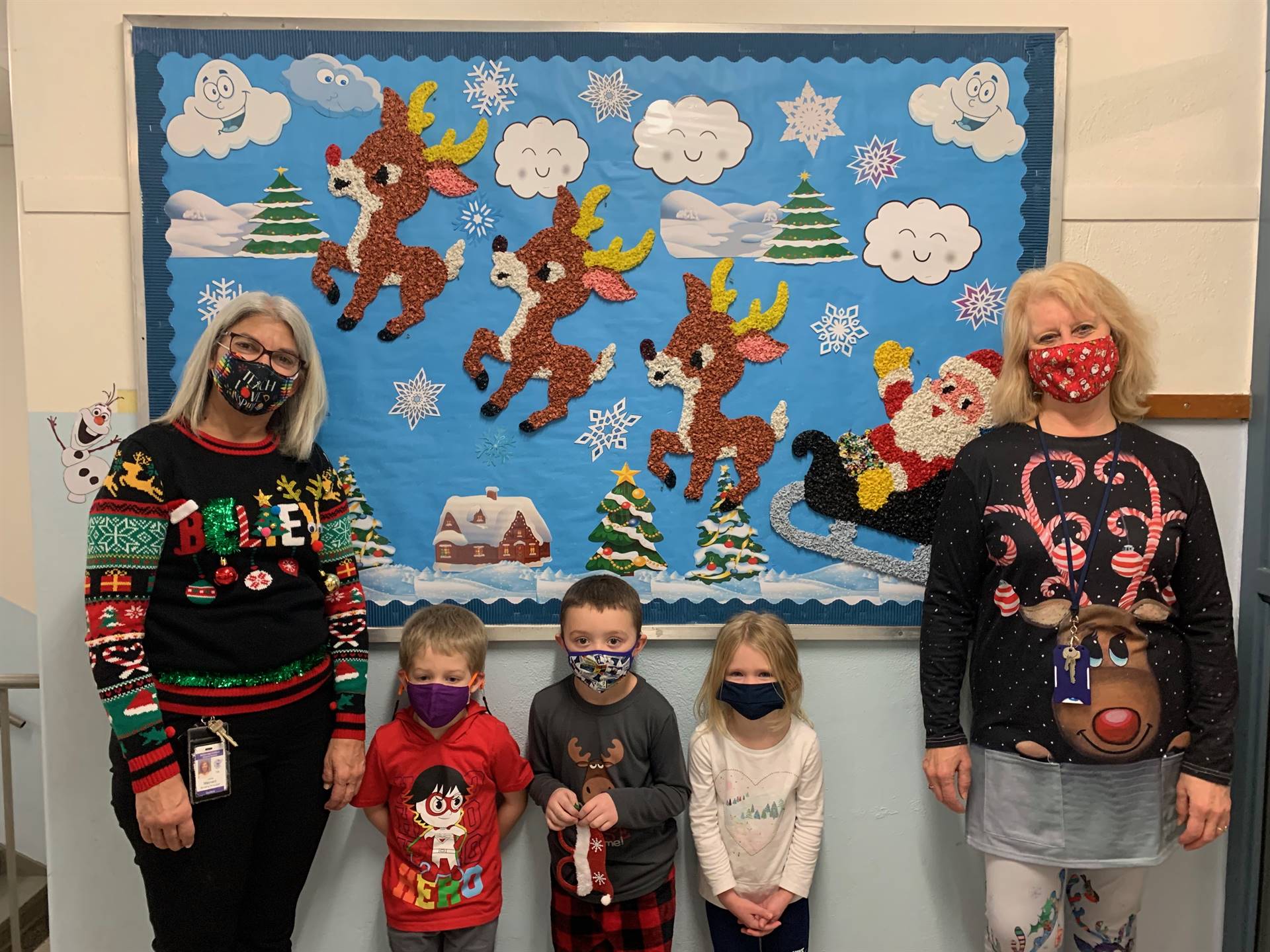 2 adults and 3 kids stand in front  bulletin board background of santa and reindeer.