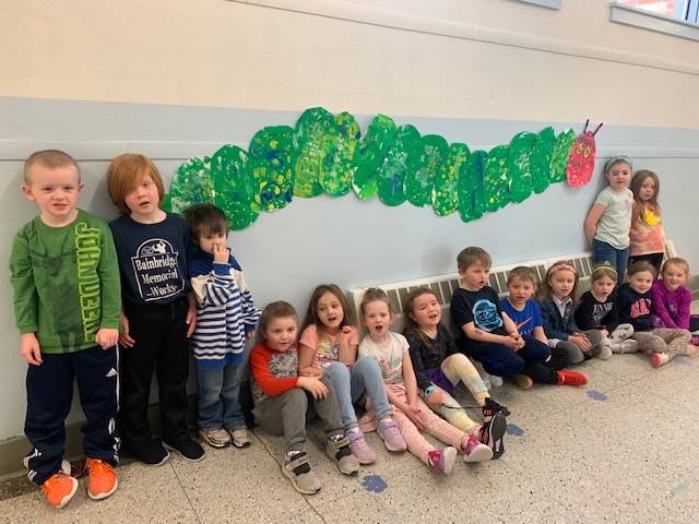 students sitting and standing in front of a giant painted caterpillar hanging on wall.
