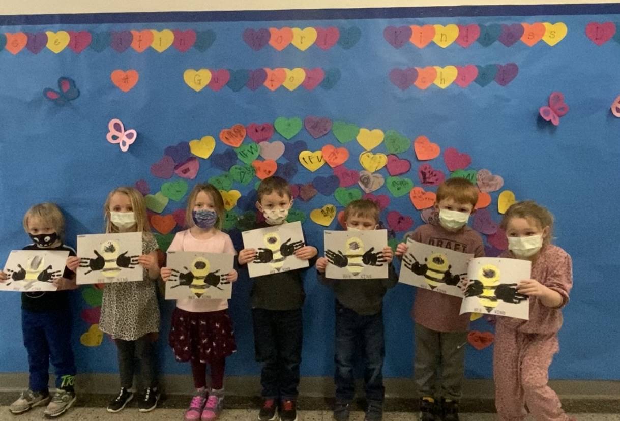 students in front of bulletin board  that is a rainbow of hearts. students are holding a bee craft