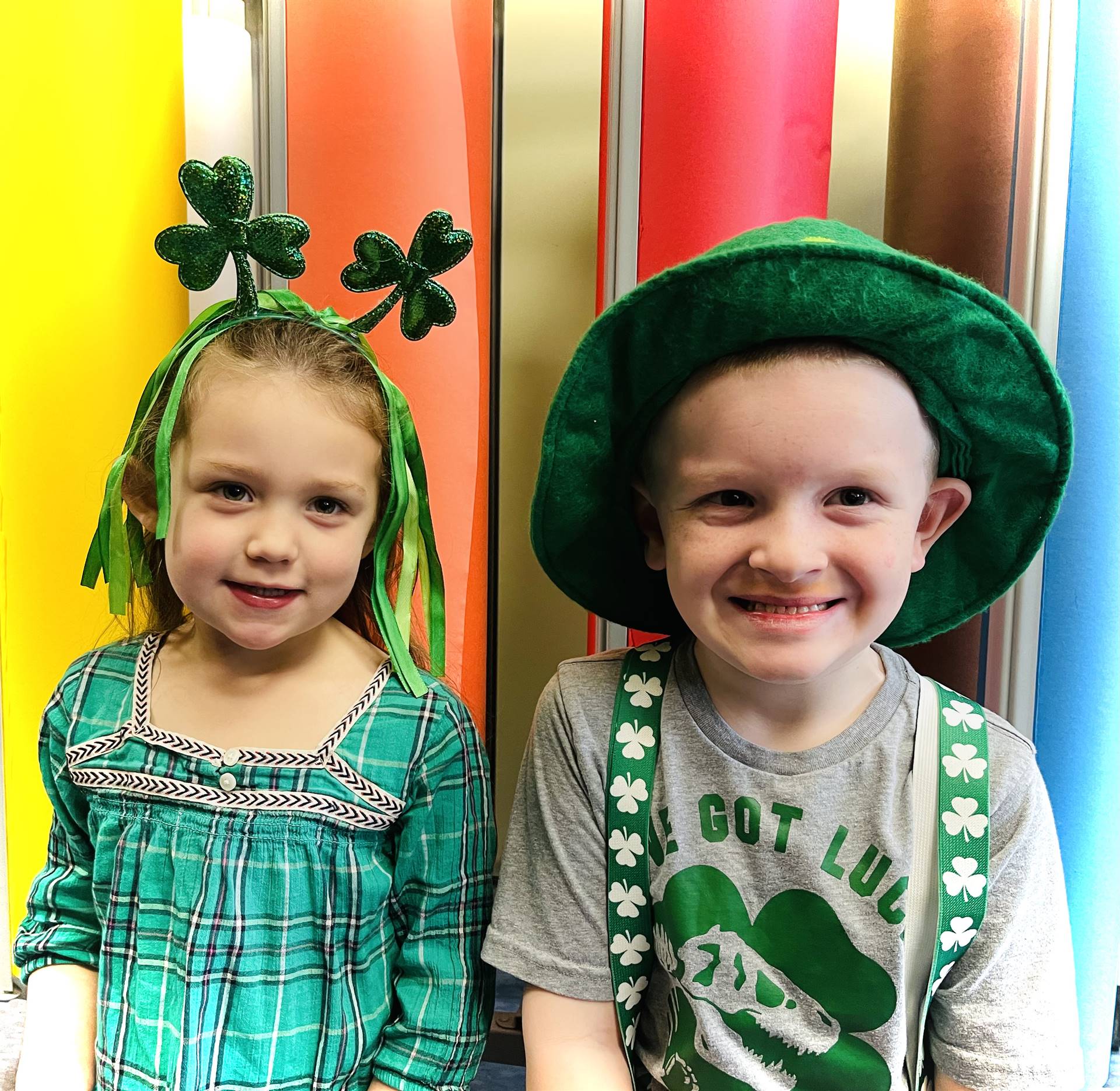 a student with a green felt hat and 1 student with a green shamrock head band against a rainbow back