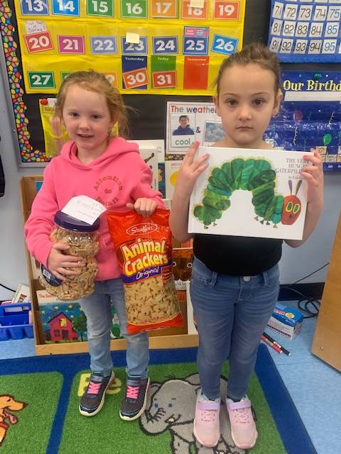 2 students. 1 holding animal crackers. 1 holding a book with a giant green caterpillar on cover.
