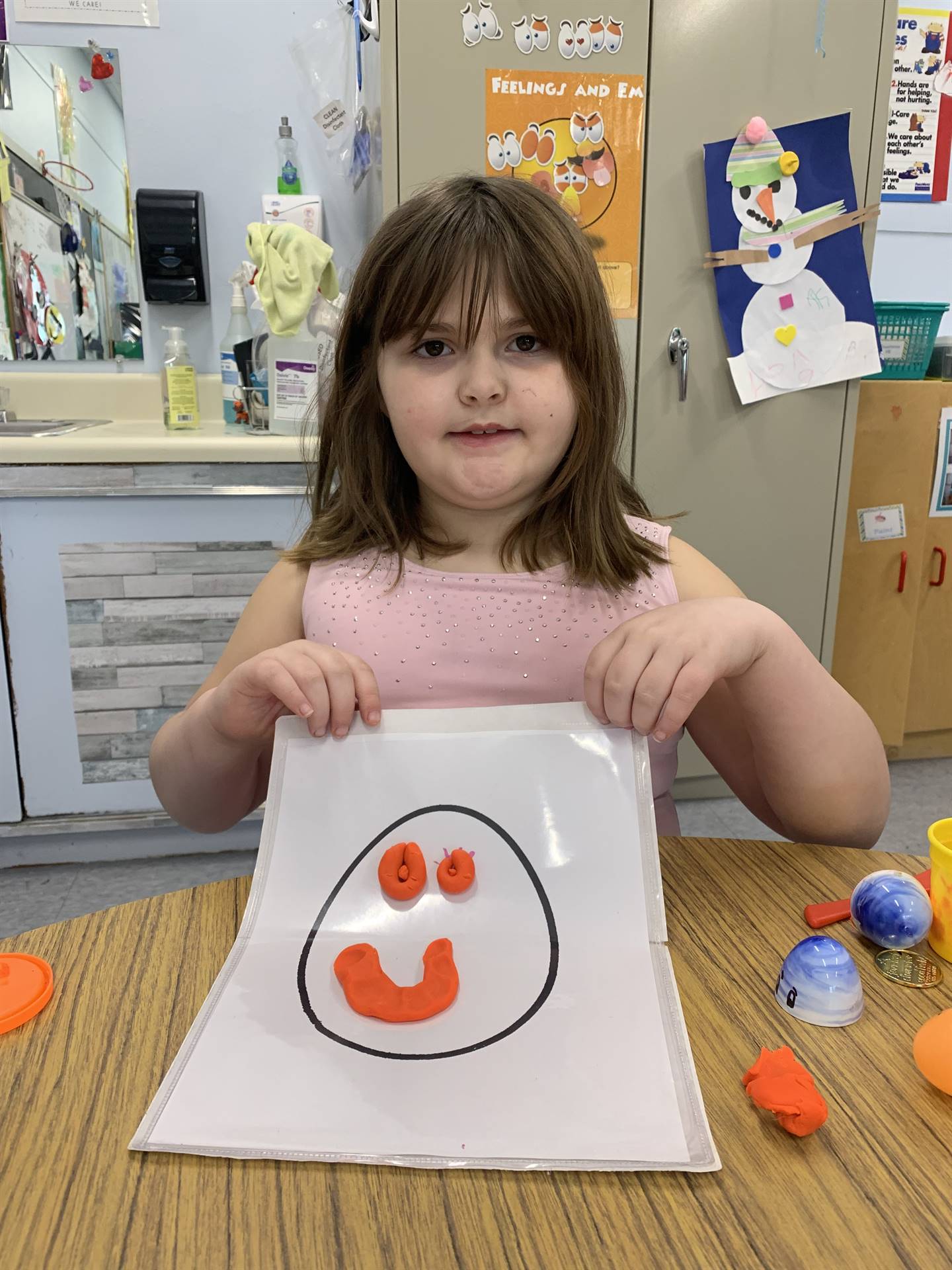student holding a paper egg shape with a playdo happy face 