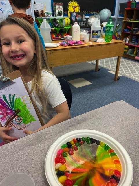 student holds up a colored rainbow and a plate of skittles melted into a rainbow