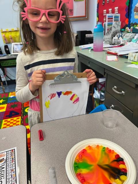 student holds up a colored rainbow and a plate of skittles melted into a rainbow