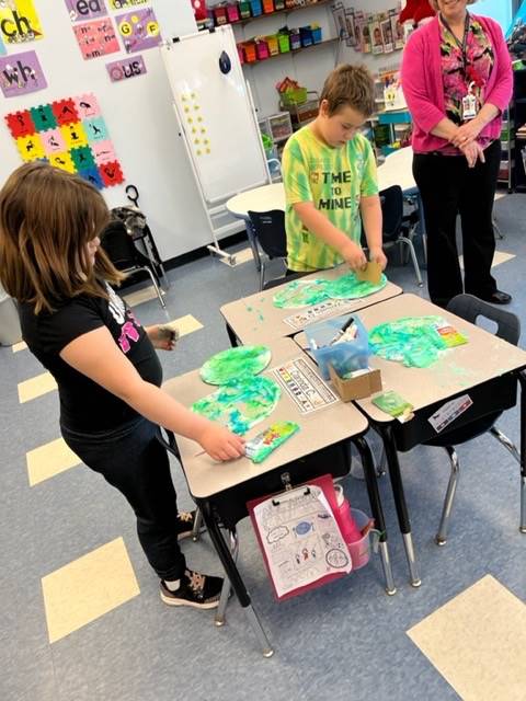  students sitting at desks with green and blue art supplies 