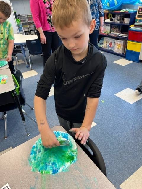 a student scrapes of blue and green shaving cream from a paper plate