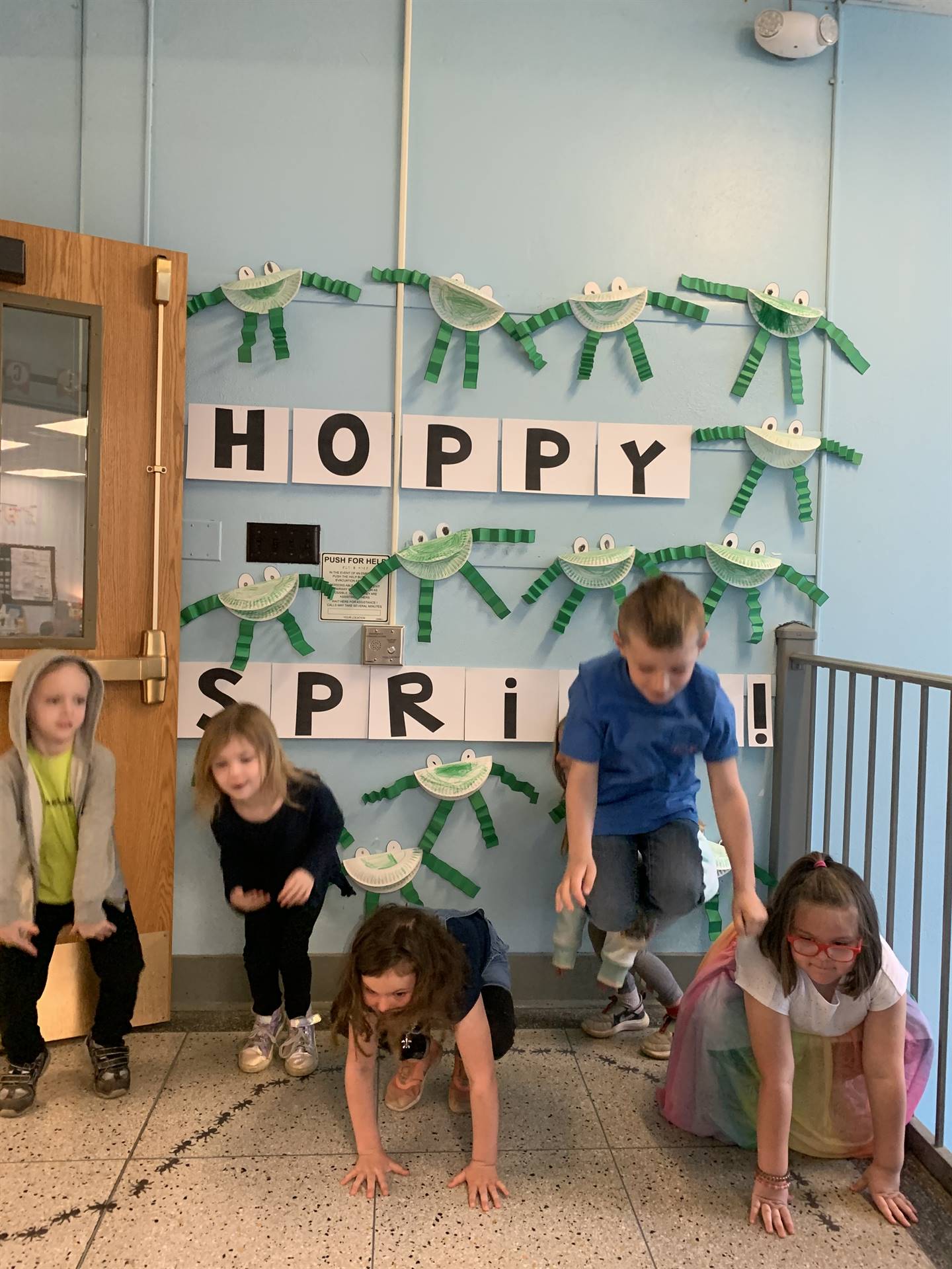 5 students hopping like frogs. background is sign "hoppy spring" with paper craft frogs.