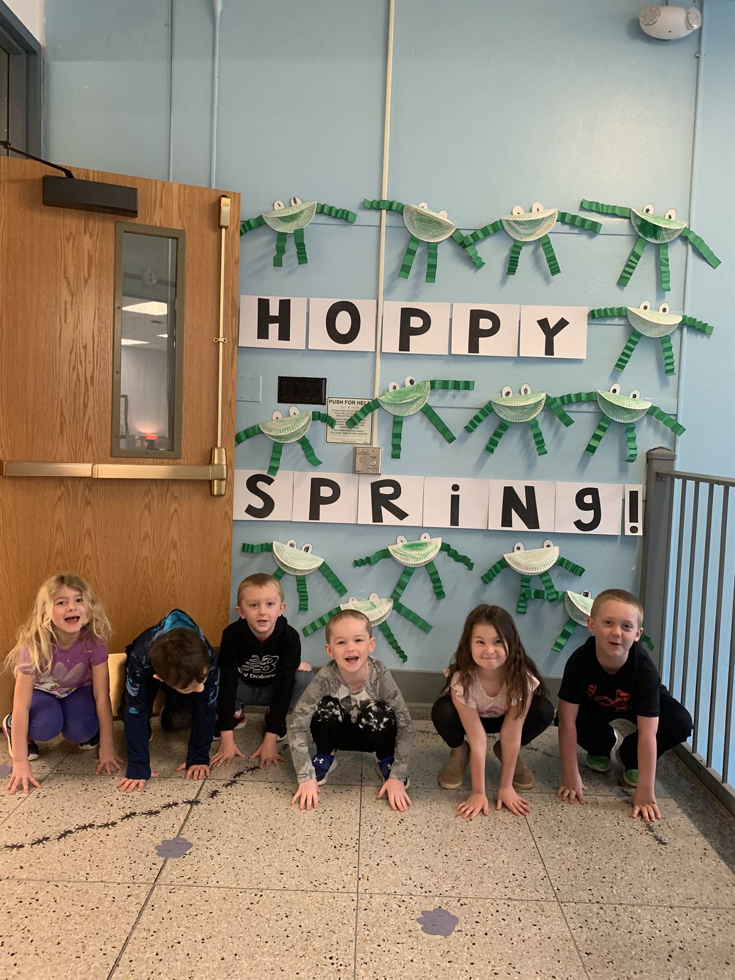 6 students hopping like frogs. background is sign "hoppy spring" with paper craft frogs.