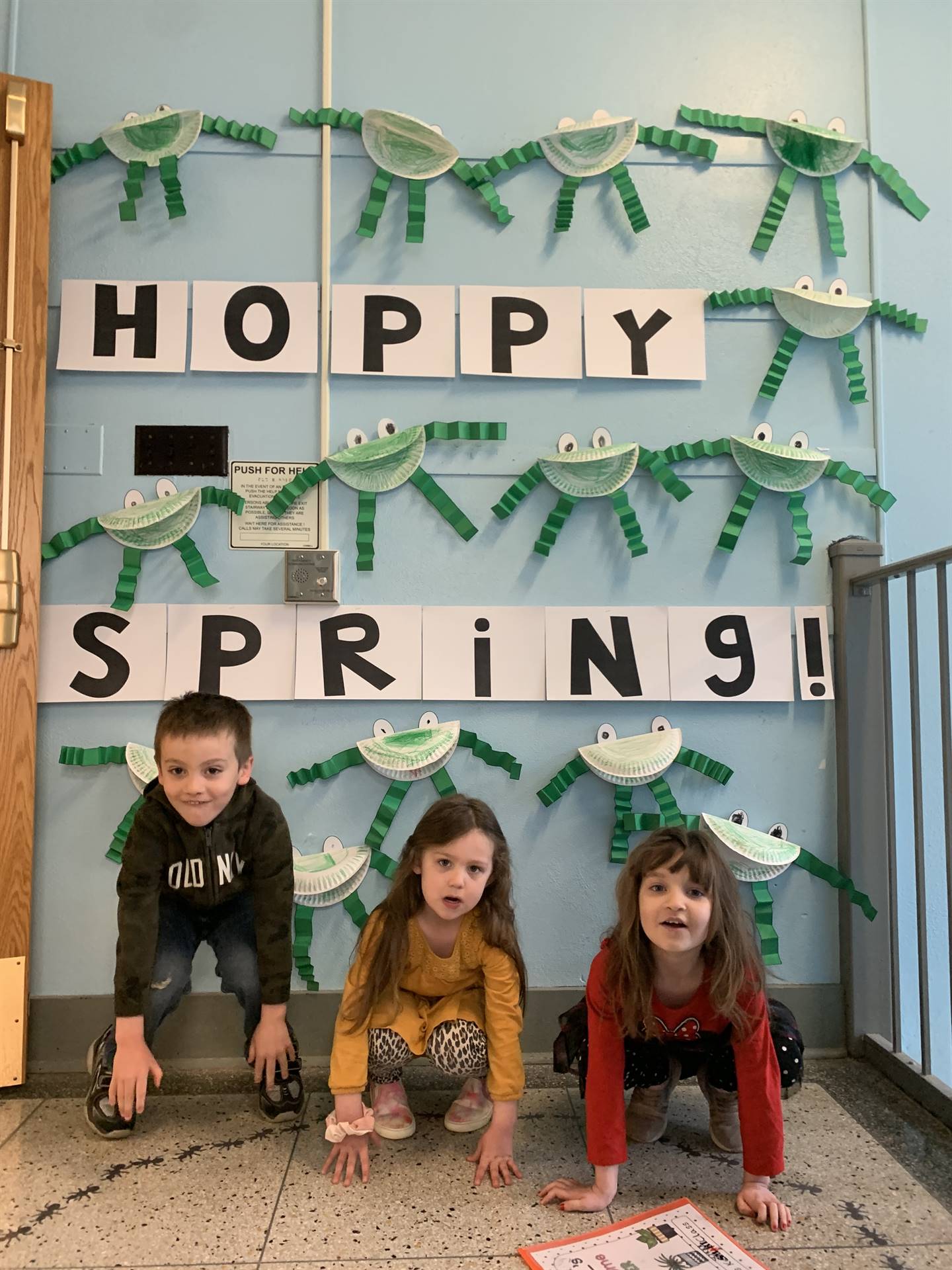 3 students hopping like frogs. background is sign "hoppy spring" with paper craft frogs.