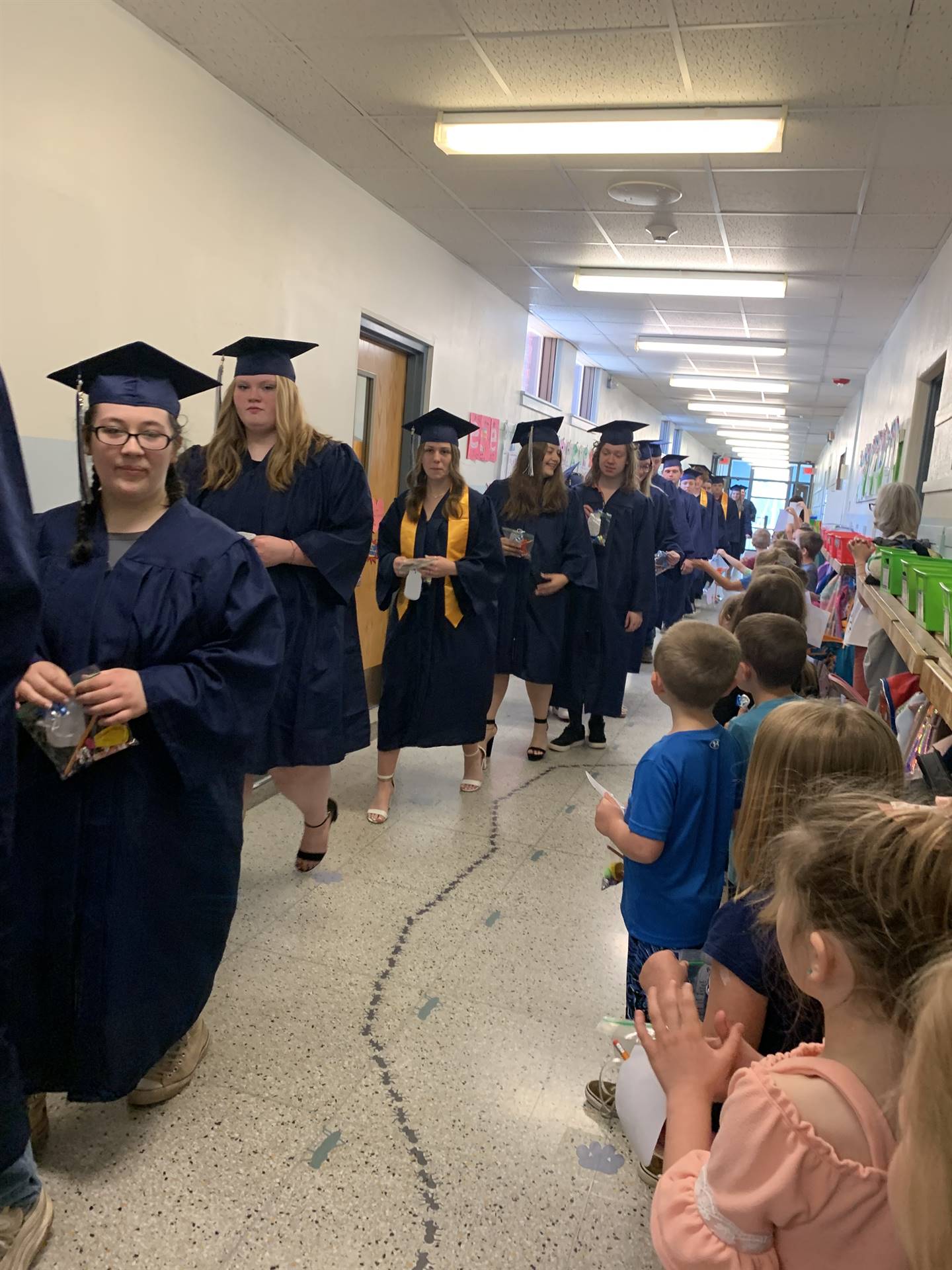 high school graduates walk in hallway while primary students 