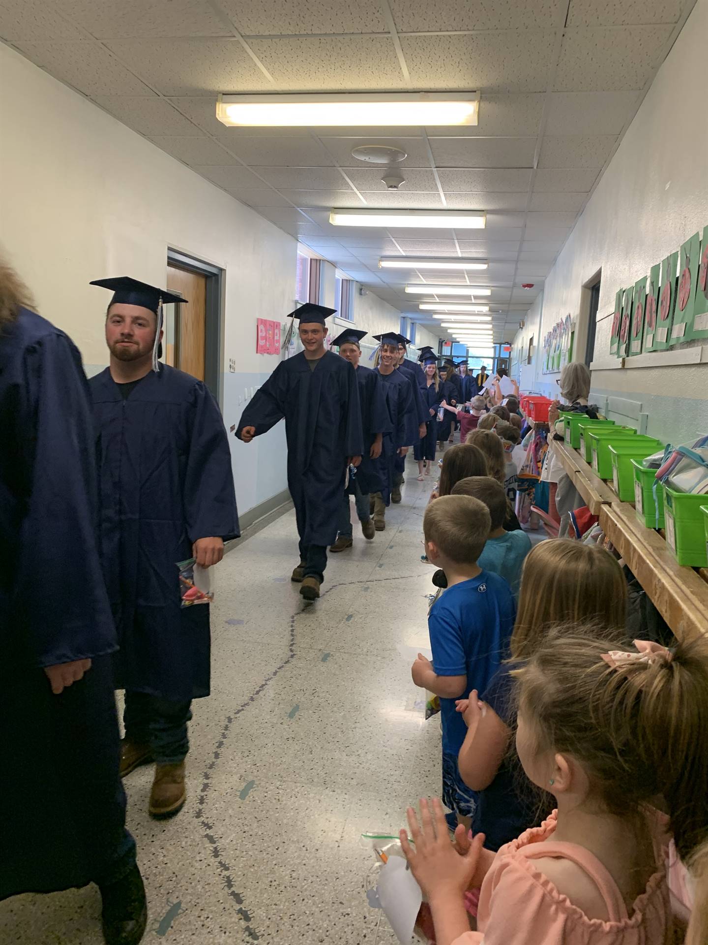 high school graduates walk in hallway while primary students clap.