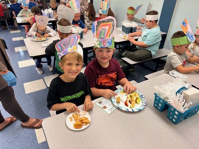 2 students eat food off a play and wear paper chef hats.