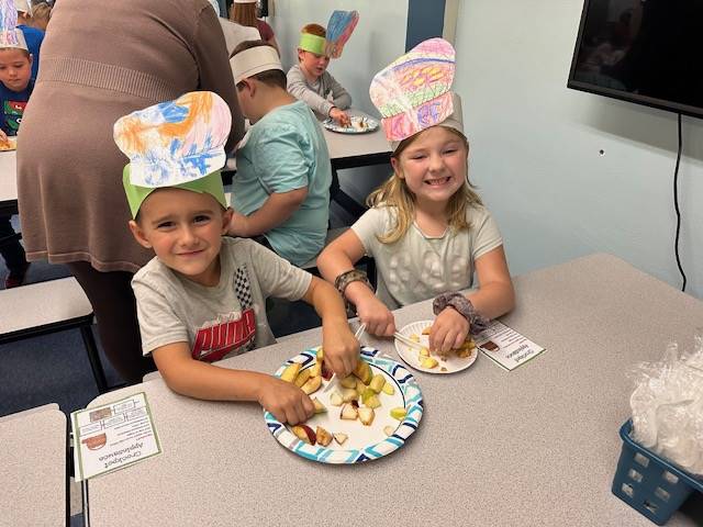2 students eat food off a play and wear paper chef hats.