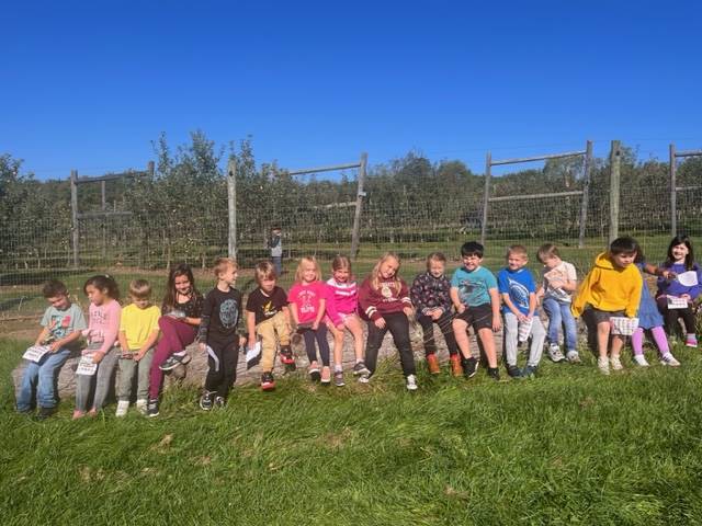a student class sits on a rock with apple orchard behind them 