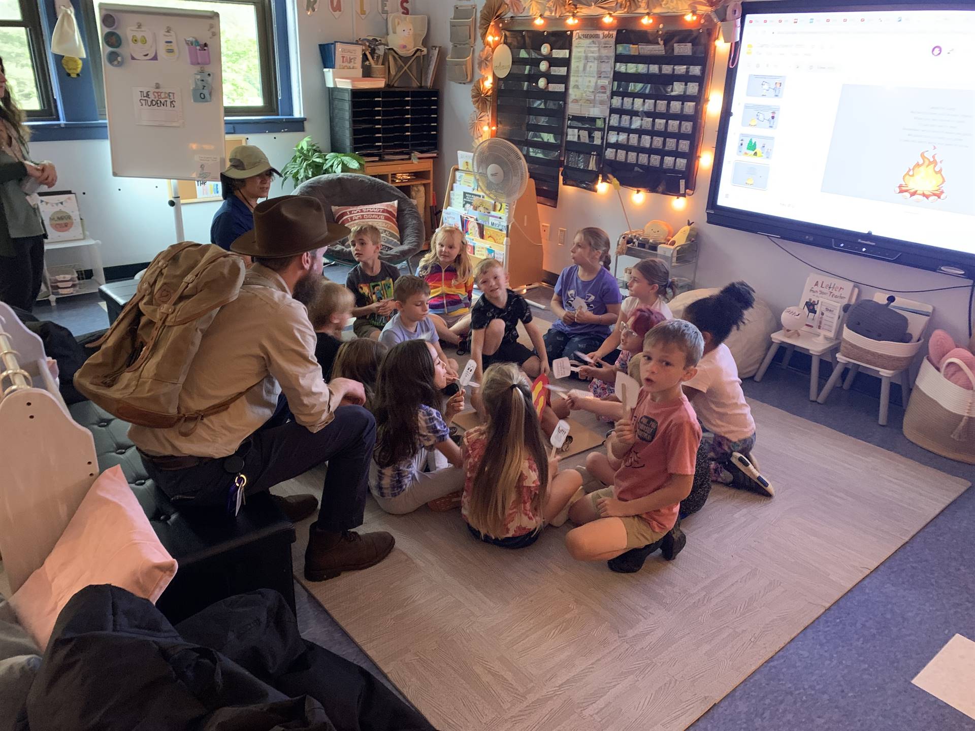 Adults dressed as campers and students sitting around a fake campfire roasting fake marshmallows