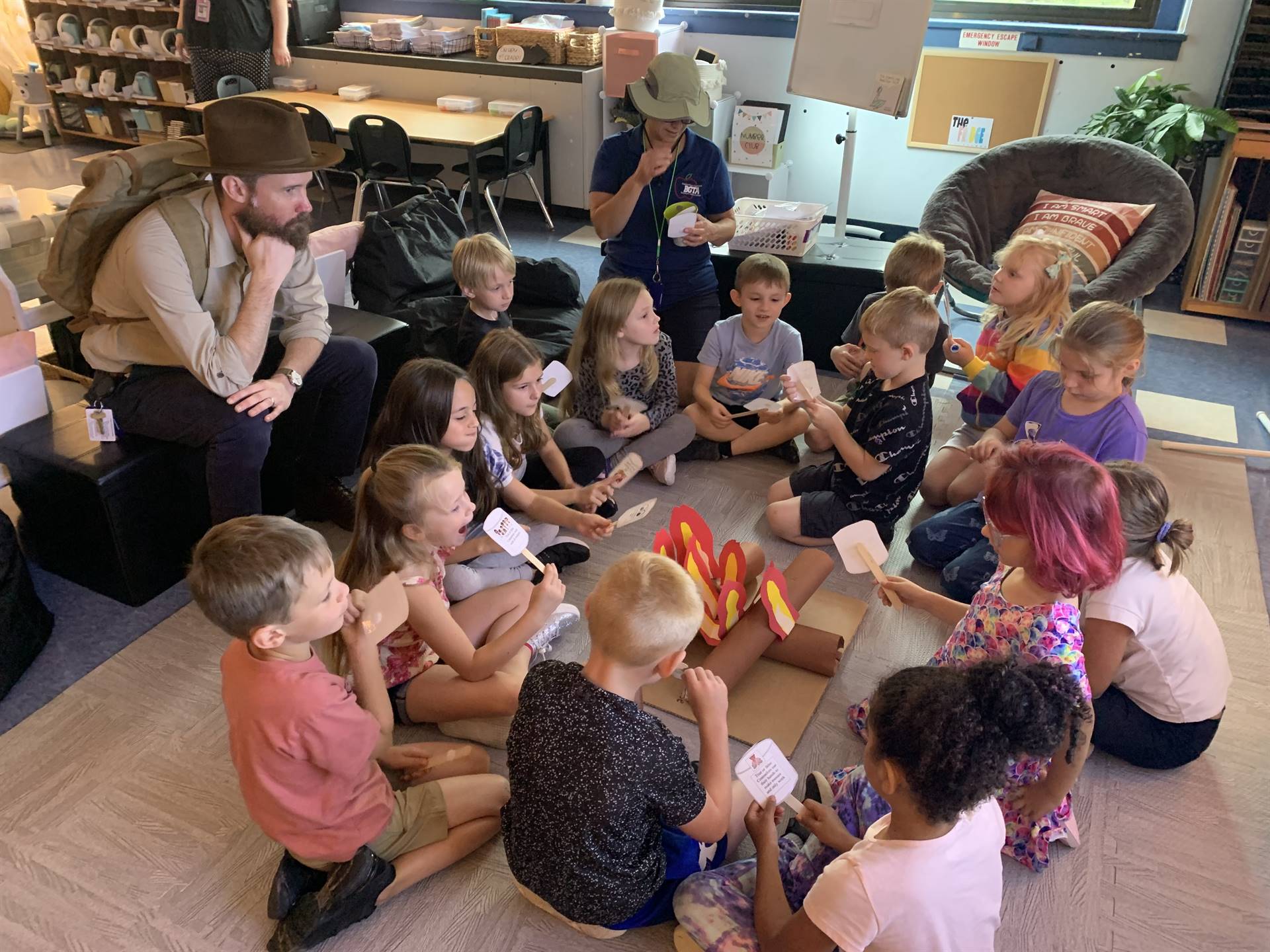 Adults dressed as campers and students sitting around a fake campfire roasting fake marshmallows