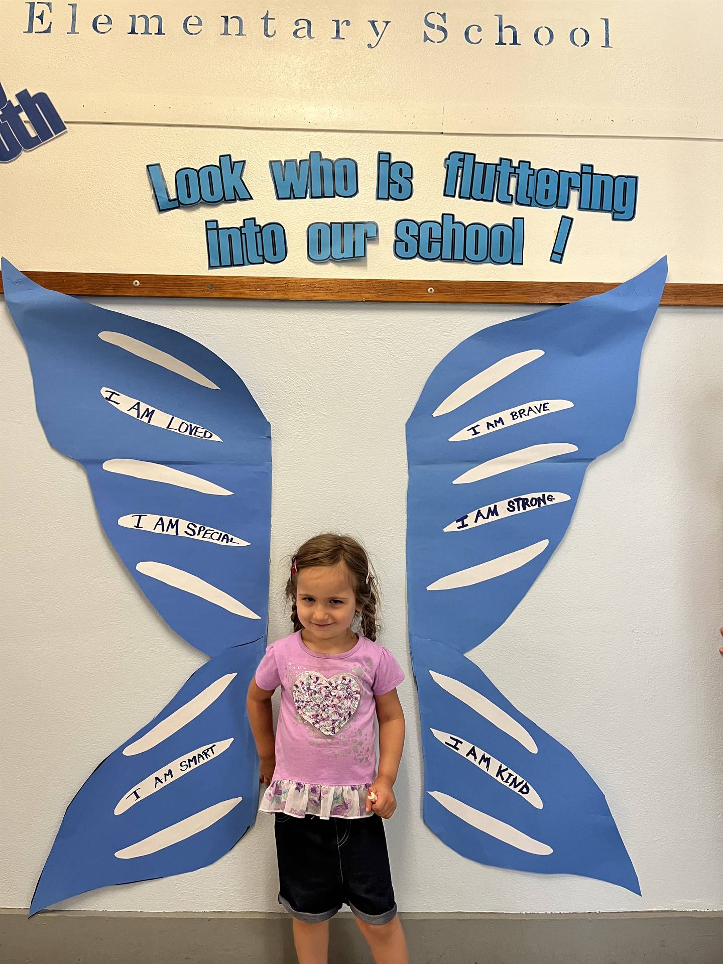 student standing between butterfly wings with sign on top saying "look who is fluttering into our sc