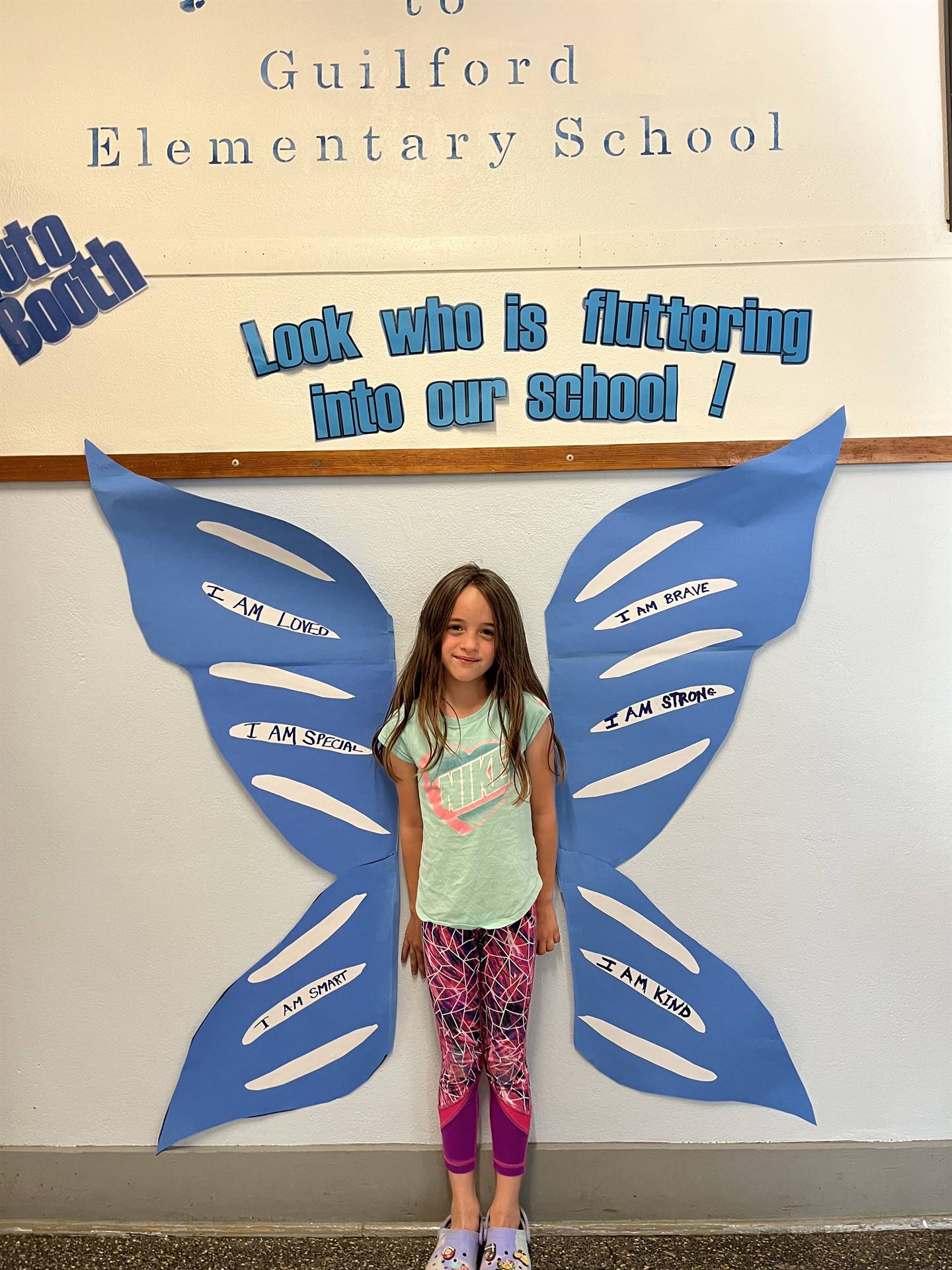 student standing between butterfly wings with sign on top saying "look who is fluttering into our sc