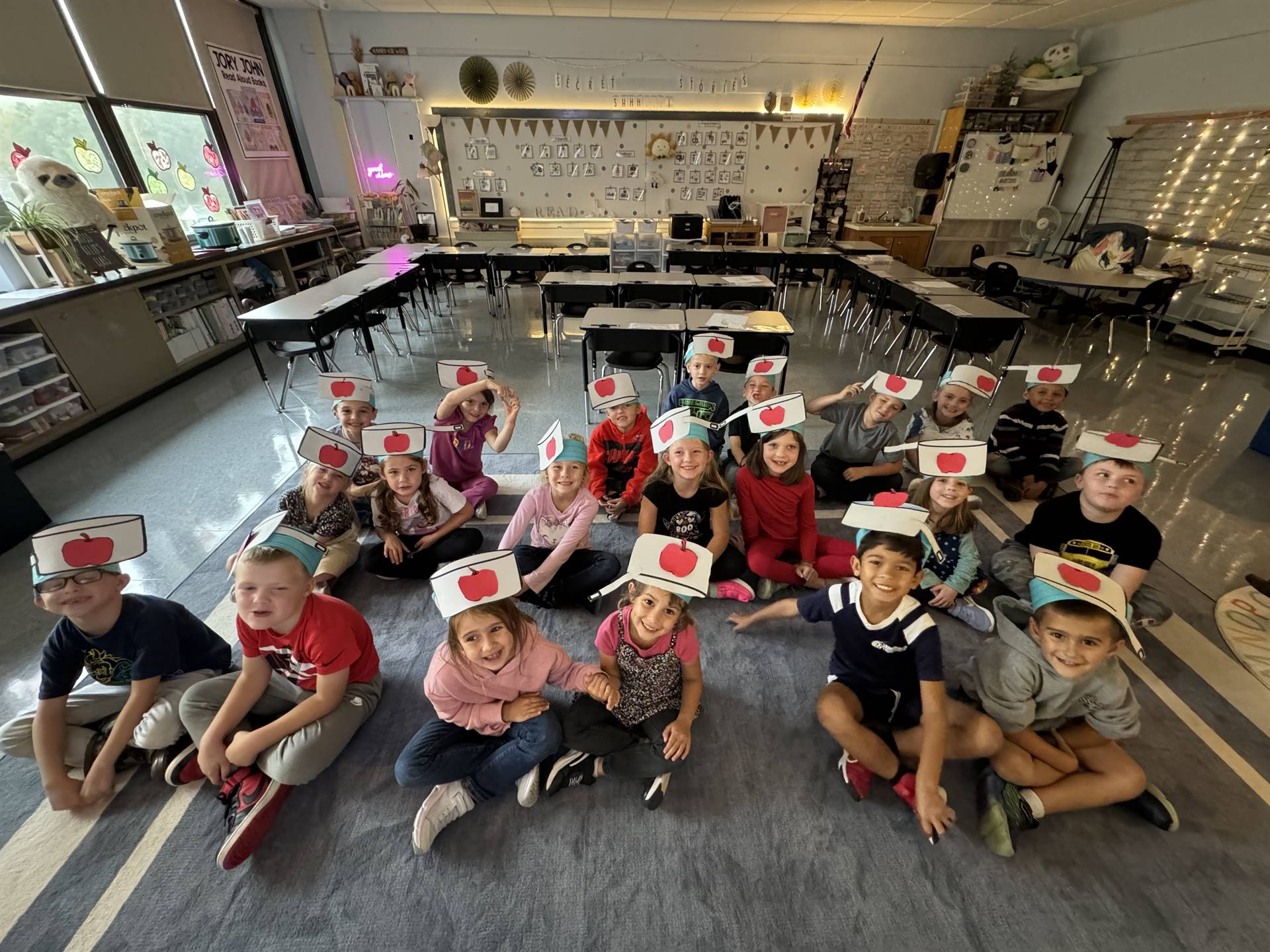 students sitting on floor wearing paper hats with red apples on the hat.