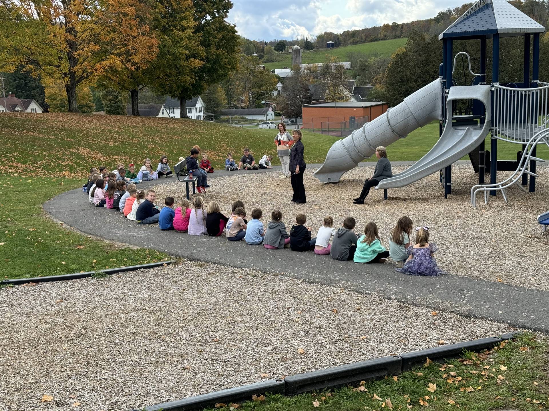 students seated outside on playground while adult speaks.