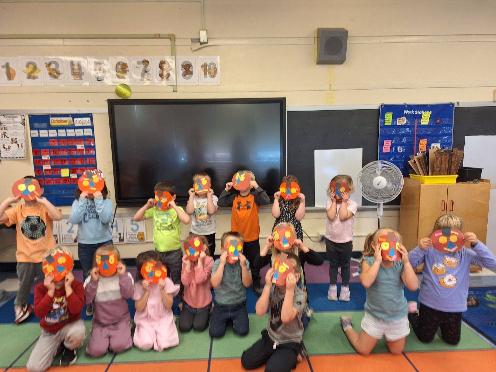 students holding up orange jackolanterns made out of paper to their faces. 