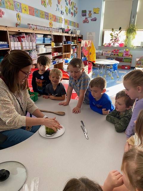 students gathered around a table watching a teacher cut an apple into sections.
