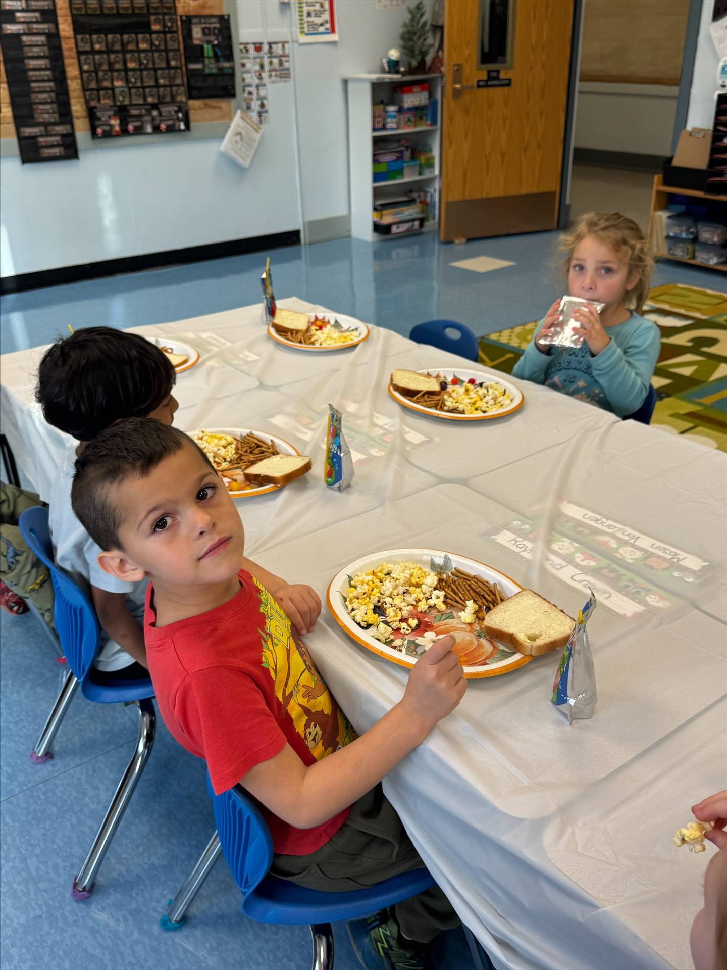 children sitting at a long white covered table with plates of food.