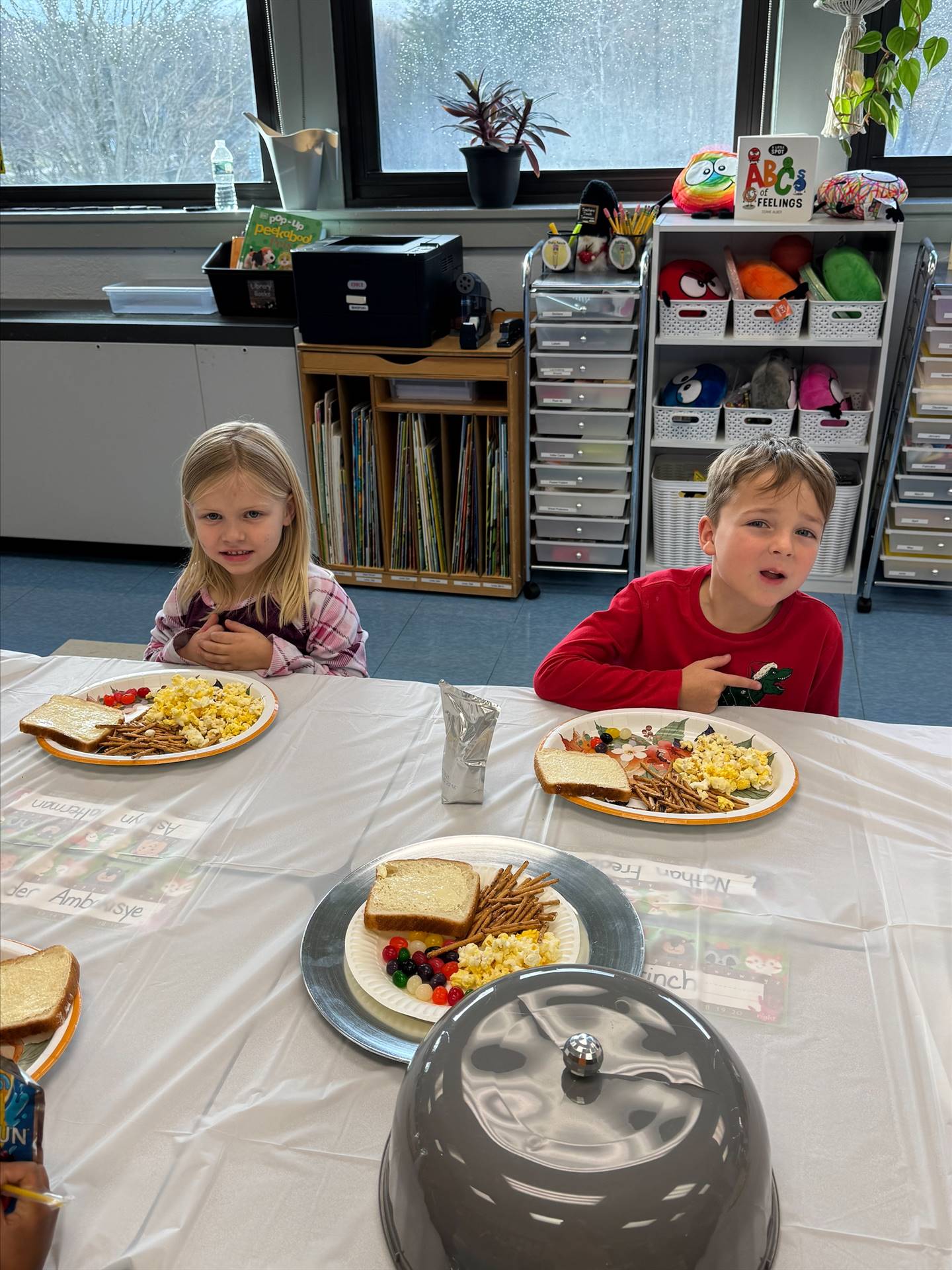 children sitting at a long white covered table with plates of food.