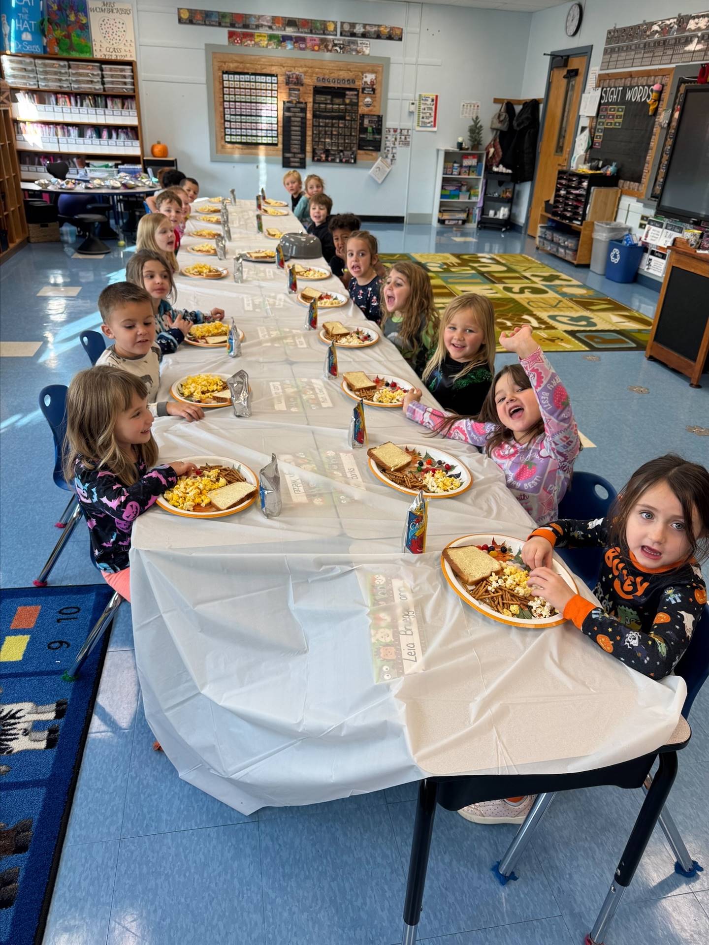 children sitting at a long white covered table with plates of food.