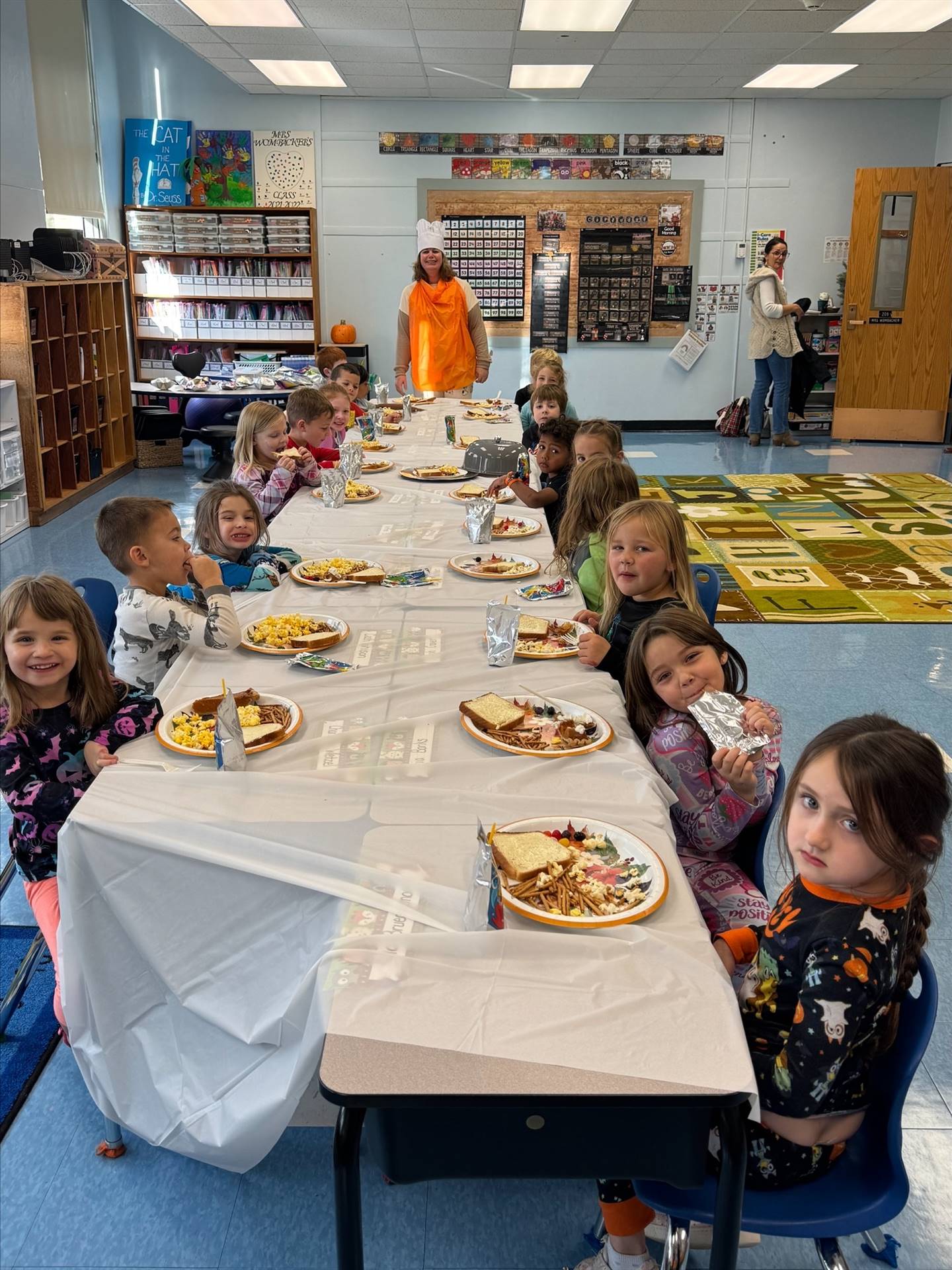 children sitting at a long white covered table with plates of food.