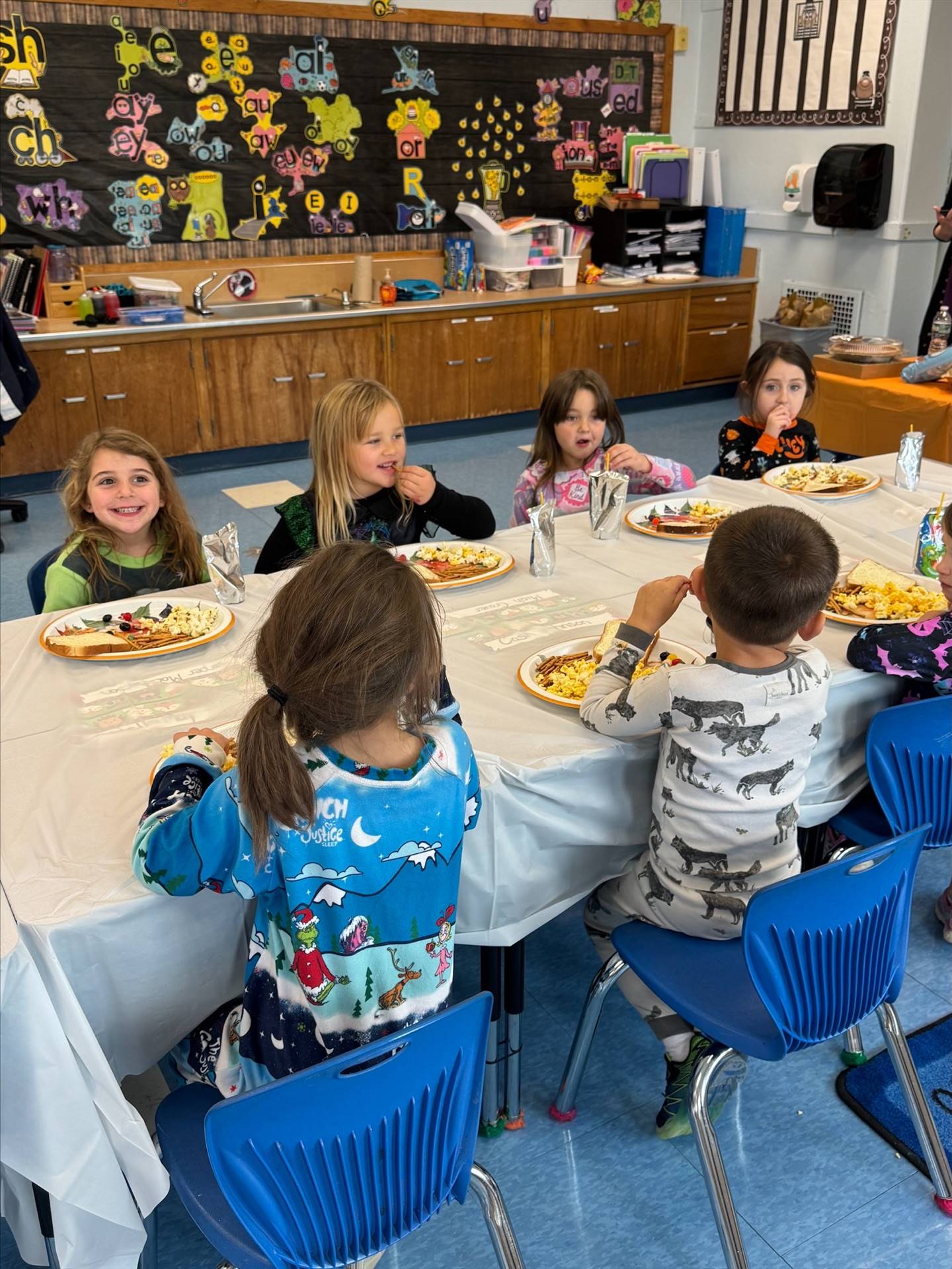 children sitting at a long white covered table with plates of food.