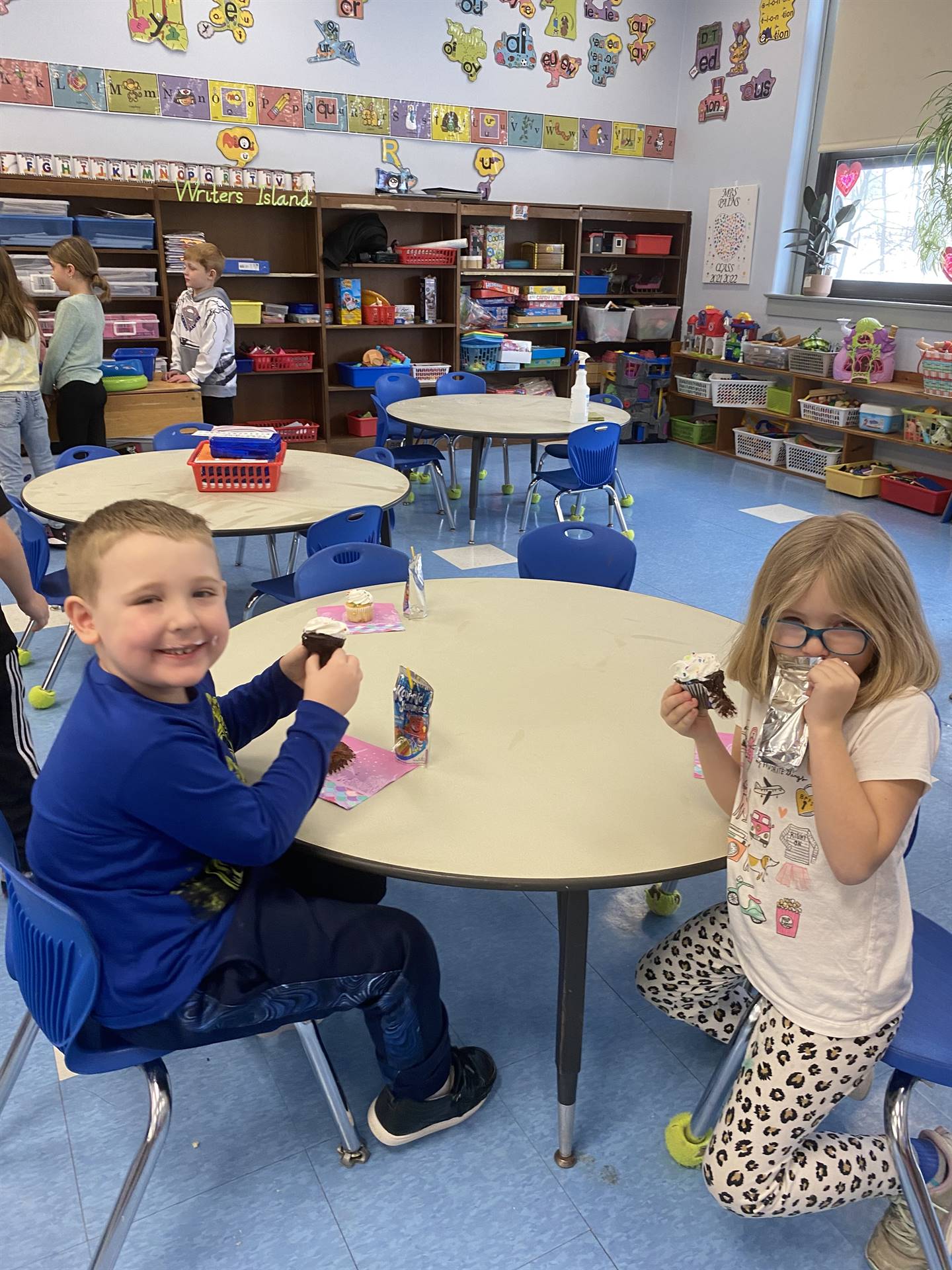 2 students sitting at a table eating cookies.