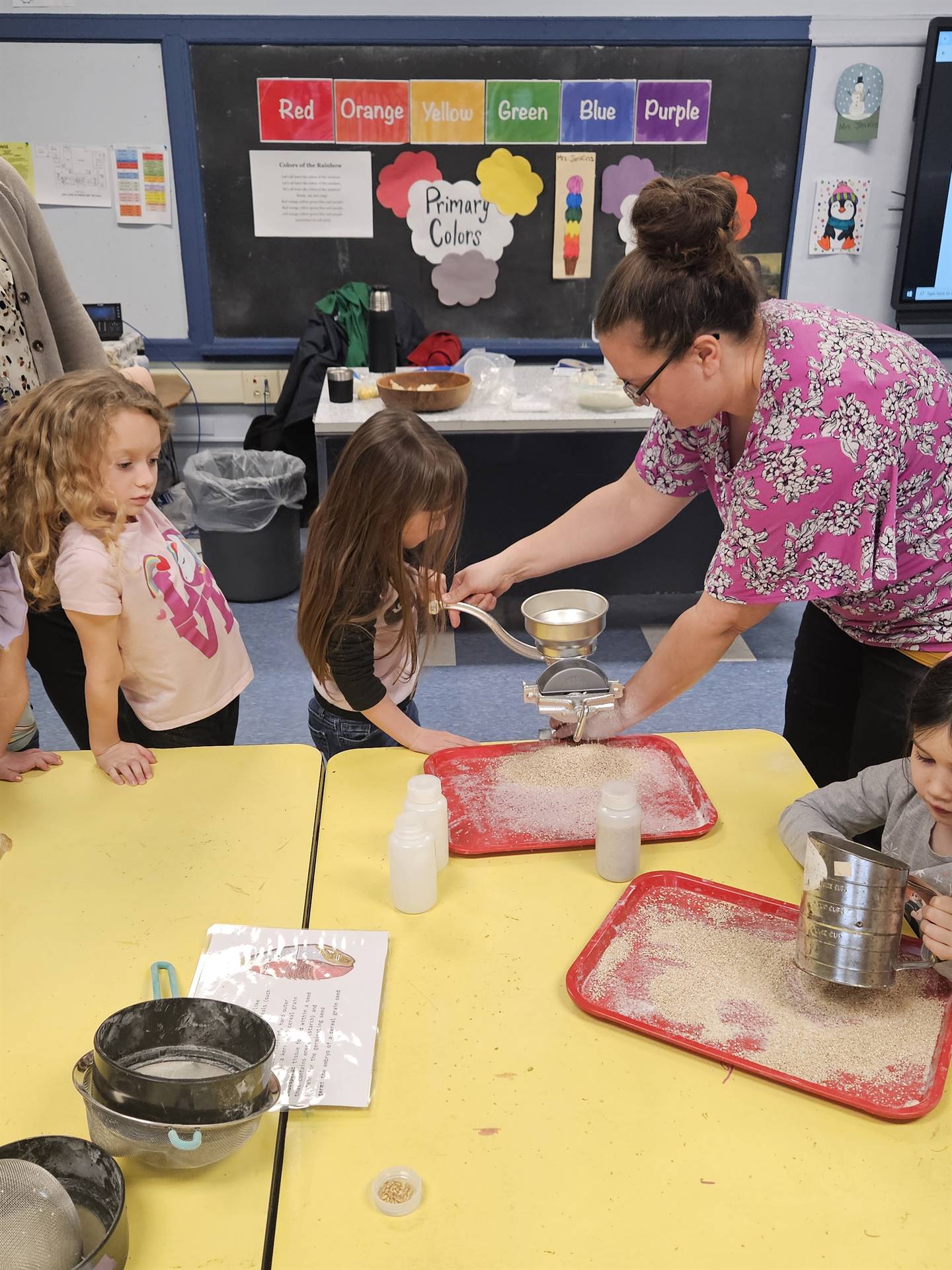 an adult helps a student use a grinder for wheat seeds.