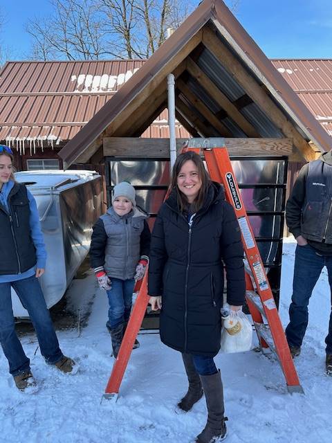 students with adults on a ladder to look into a maple syrup sap collecting bin.