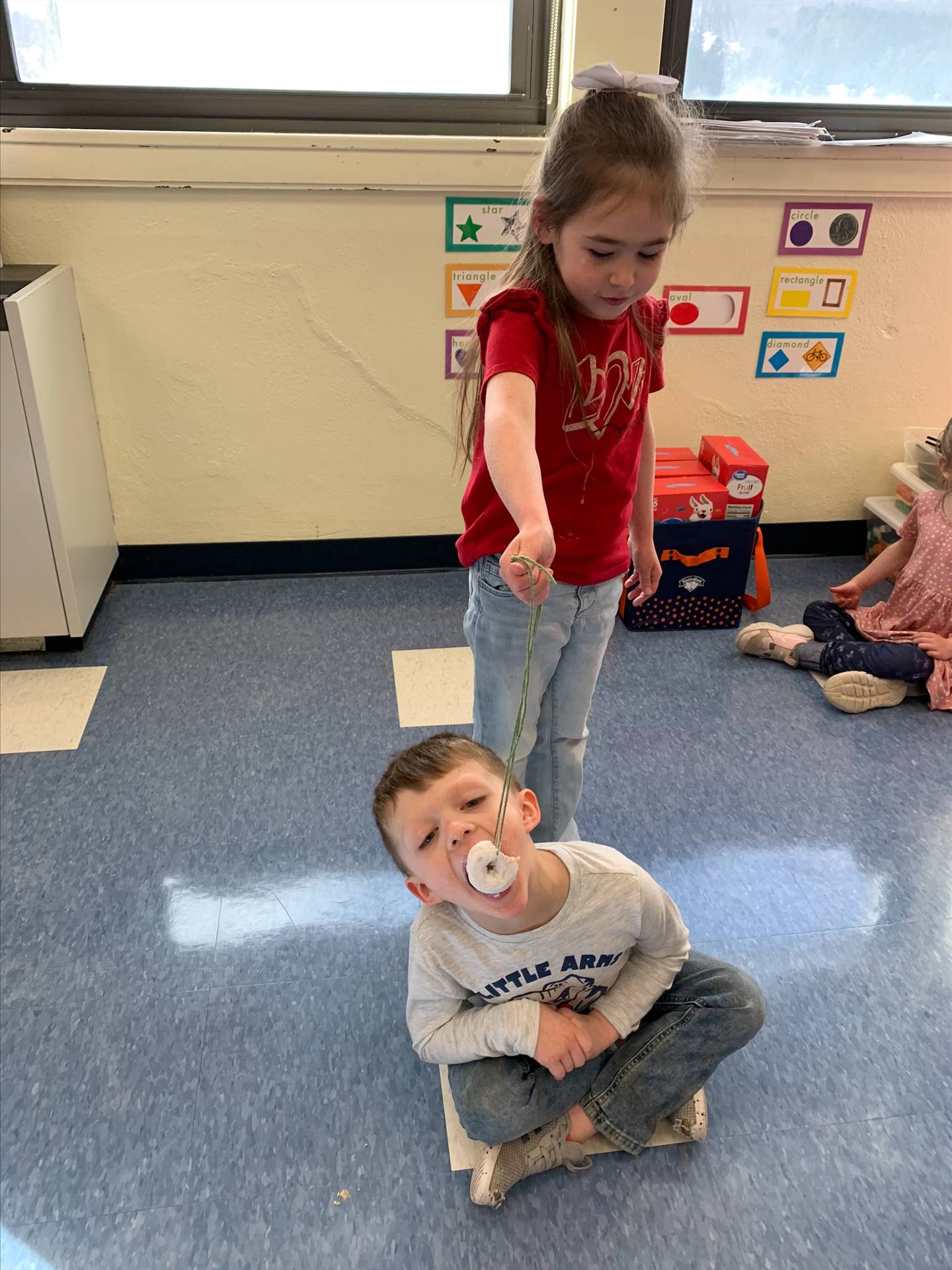 standing student hold donut on string while sitting student tries to bite it off.