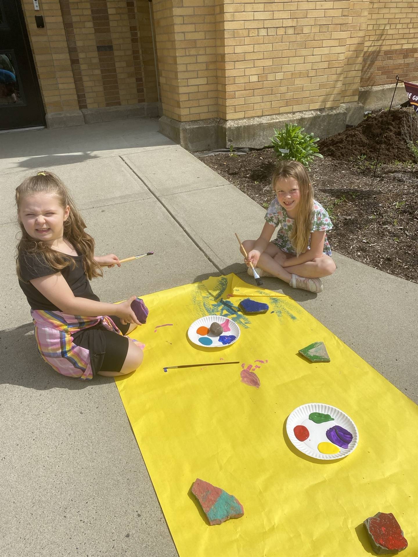 students on sidewalk painting rocks on a yellow piece of paper.