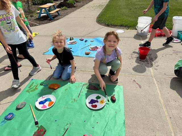 students on sidewalk painting rocks on a green  piece of paper.